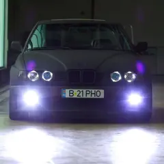 Matte-black BMW 7-series on E30 M3 body kit, close front portrait with LED angel-eye halo headlights glowing in a workshop