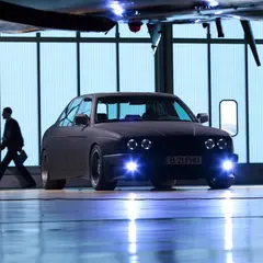 Matte-black BMW 7-series with an E30 M3 body kit, front view with LED angel-eye headlights lit up inside an aircraft hangar